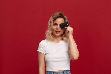 Portrait of a girl with curly blond hair in a white t-shirt standing on a red background. Model smiles at the camera and holds bank card covering half of a face.