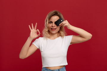 Portrait of a girl with curly blond hair in a white t-shirt standing on a red background. Model smiles at the camera, holds bank card covering half of a face and showing gesture of OK-symbol.