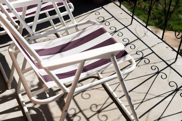 Close up of folding chair on concrete balcony floor with shadows of metal balcony fence. Relaxing on the sun in summer time