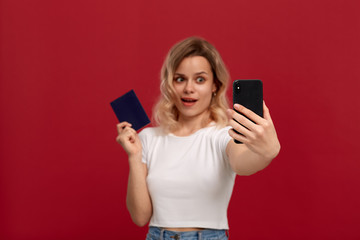 Fototapeta premium Portrait of a girl with curly blond hair in a white t-shirt on a red background. Happy model makes selfie with the mobile phone holding passport