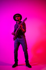 Obraz na płótnie Canvas Young african-american musician playing the guitar like a rockstar on gradient purple-pink background in neon light. Concept of music, hobby. Joyful attractive guy improvising and singing a song.