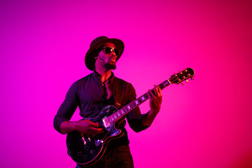 Obraz na płótnie Canvas Young african-american musician playing the guitar like a rockstar on gradient purple-pink background in neon light. Concept of music, hobby. Joyful attractive guy improvising. Colorful portrait.