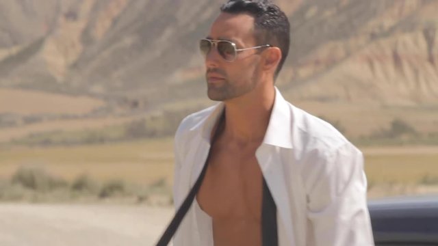 Handsome businessman in white shirt open opening the door, coming out of his sports car, then standing and alone in the desert and removing sunglasses. Beautiful abs, fitness model, mountains.