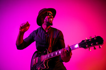 Obraz premium Young african-american musician playing the guitar like a rockstar on gradient purple-pink background in neon light. Concept of music, hobby. Joyful attractive guy improvising and singing a song.