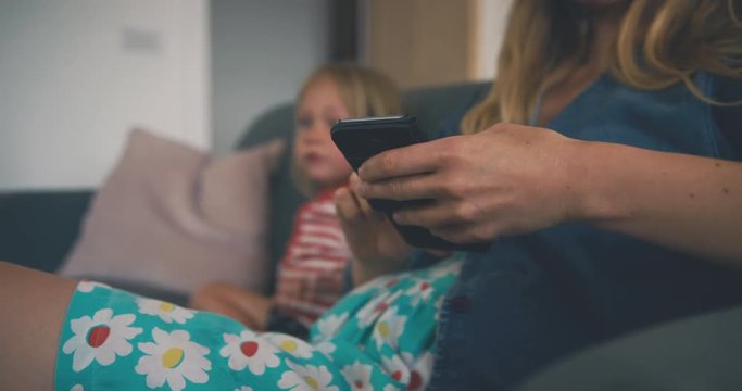 Mother using smartphone on sofa with toddler watching television