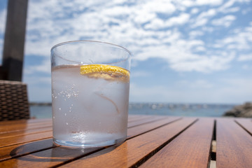 A glass of cold sparkly water with a lemon slice on a wooden table in the beach