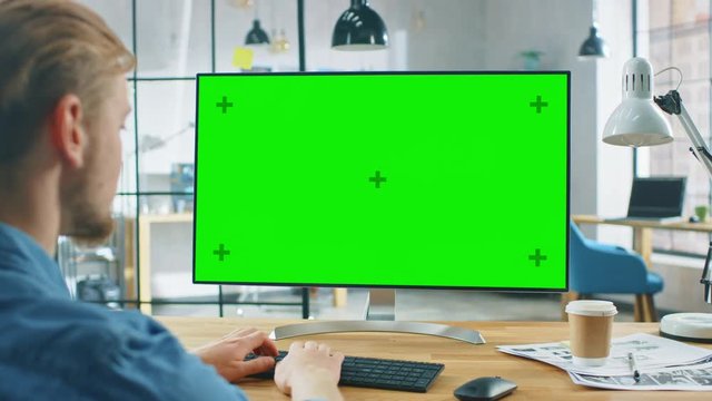 Over the Shoulder Shot: Man Uses Laptop Computer with Green Mock-up Screen while Sitting at His Desk. In the Background Stylish Modern Open Space Creative Business Office
