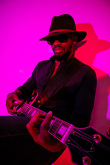 Obraz premium Young african-american musician playing the guitar like a rockstar on gradient purple-pink background in neon light. Concept of music, hobby. Joyful attractive guy improvising. Colorful portrait.