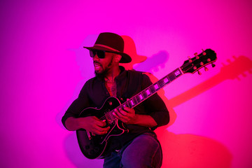 Obraz premium Young african-american musician playing the guitar like a rockstar on gradient purple-pink background in neon light. Concept of music, hobby. Joyful attractive guy improvising. Colorful portrait.
