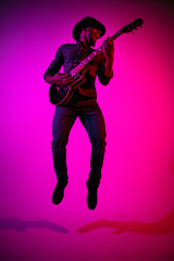 Fototapeta na wymiar Young african-american musician playing the guitar like a rockstar on gradient purple-pink background in neon light. Concept of music, hobby. Joyful attractive guy improvising. Colorful portrait.