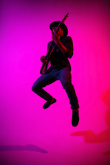 Fototapeta na wymiar Young african-american musician playing the guitar like a rockstar on gradient purple-pink background in neon light. Concept of music, hobby. Joyful attractive guy improvising. Colorful portrait.