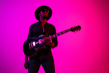 Obraz premium Young african-american musician playing the guitar like a rockstar on gradient purple-pink background in neon light. Concept of music, hobby. Joyful attractive guy improvising. Colorful portrait.
