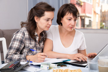Two smiling women sitting at home table with laptop