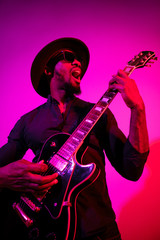 Plakat Young african-american musician playing the guitar like a rockstar on gradient purple-pink background in neon light. Concept of music, hobby. Joyful attractive guy improvising. Colorful portrait.