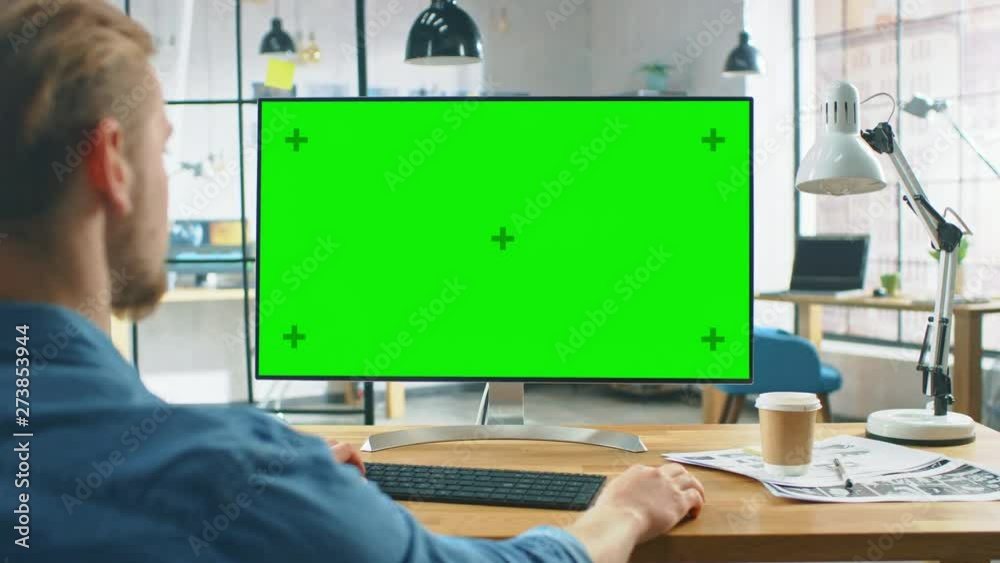 Poster over the shoulder shot: man uses laptop computer with green mock-up screen while sitting at his desk