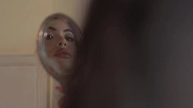 Attractive brunette Arab woman holding Old Silver makeup mirror vintage, looking at her reflection in the mirror, touching her lips. Hands in red varnish. Hotel room in Paris with Sun light flare. 
