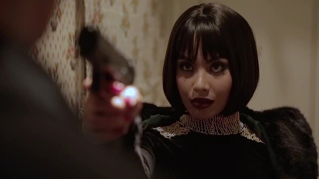 Sexy brunette Arab woman in tight black dress with jewels and big pearl necklace pointing a gun on her husband who cheated on her in a retro hotel with walls adorned with floral tapestry. Close Up.