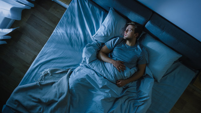 Top View Of Handsome Young Man Sleeping Cozily On A Bed In His Bedroom At Night. Blue Nightly Colors With Cold Weak Lamppost Light Shining Through The Window.