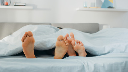 Cute Male and Female Lying in Bed with Feet Sticking from Under the Blanket, Touching, Flirting, Fooling Around. Bright and Cozy Bedroom.