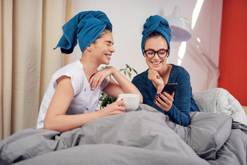 Obraz na płótnie Canvas Two beautiful Caucasian girlfriends with towels on heads sitting on bed in bedroom, drinking coffee and gossiping.