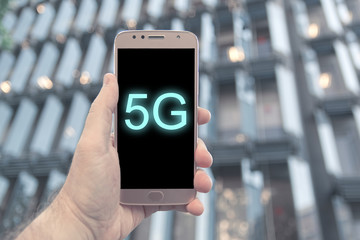 Hand holding a 5G compatible mobile smartphone against a modern building facade in Barelona. Empty copy space for Editor's text.