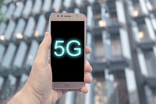 Hand holding a 5G compatible mobile smartphone against a modern building facade in Barelona. Empty copy space for Editor's text.