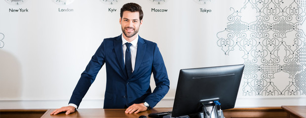 panoramic shot of happy receptionist standing at reception desk in hotel