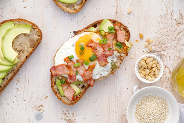 Open sandwich made of slices of sourdough bread with addition of avocado, egg, bacon, fresh scallion on a wooden white table, top view. 
