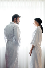 low angle view of happy man and attractive woman in bathrobes standing and looking at each other