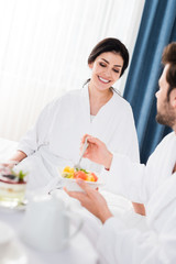 selective focus of happy woman looking at bearded man holding fork near tasty fruit salad