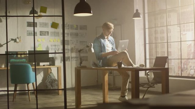 Creative Designer Walks around His Studio, Sits on His Desk Holds Laptop on the Knees while Working on the Project. Video, Smartphone Application and Gaming Content Development Freelancer
