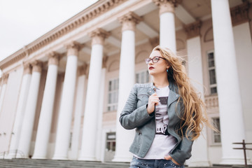 Fototapeta premium Young caucasian blondie hipster woman with long curly hairstyle in eye glasses in the city street. Freelance, freedom, beauty, happy lifestyle concept