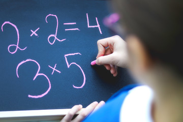 Top view of schoolgirl in glasses doing sums writing on blackboard by chalk. Back to school and child education concept. Selective focus