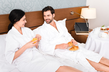 Fototapeta premium cheerful man holding croissants while looking at woman with glass of orange juice