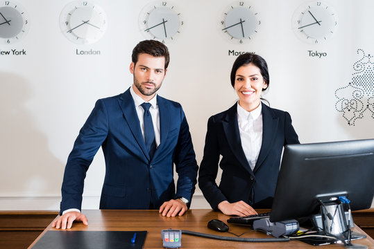 cheerful woman near serious receptionist in suit standing at reception desk