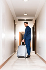 cheerful bearded man smiling while standing with suitcase in hotel corridor