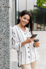 Image of brunette young woman drinking takeaway coffee and holding cellphone while standing over building