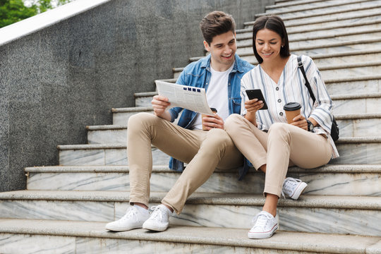 Happy Young Amazing Loving Couple Business People Colleagues Outdoors Outside On Steps Reading Newspaper Drinking Coffee.