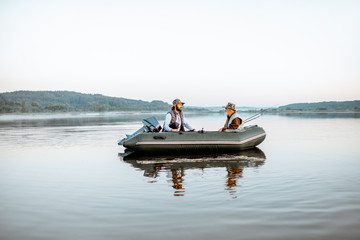 Grandfather with adult son fishing on the inflatable boat on the lake with calm water early in the morning. Wide landscape view
