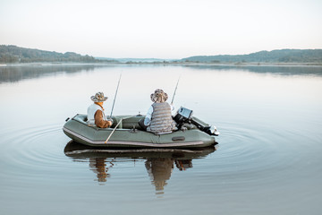 Grandfather with adult son fishing on the inflatable boat on the lake with calm water early in the...