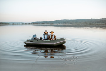 Grandfather with adult son fishing on the inflatable boat on the lake with calm water early in the...