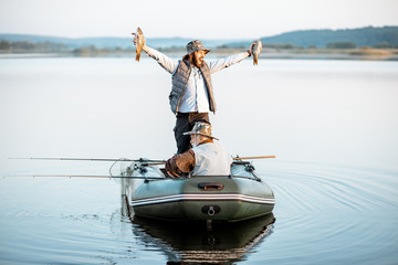Happy man holding freshly caught fish while standing on the boat with grandfather on the lake early in the morning