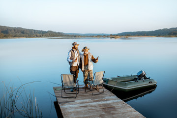 Grandfather with adult son standing together on the wooden pier, enjoying the sunrise while fishing on the lake early in the morning
