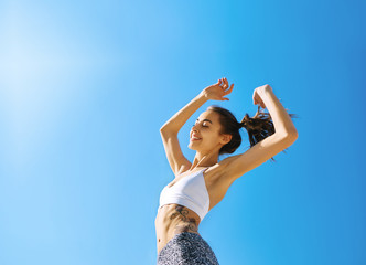 Obraz na płótnie Canvas Happy woman with tanned slim body and tattoo posing in sport wear against blue sky. Healthy smiling female enjoying sunny summer day. Summertime concept.
