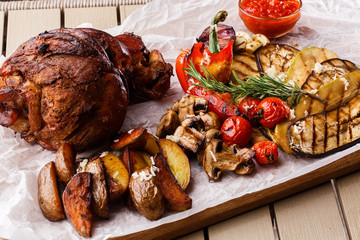 Grilled pork knuckle with grilled tomatoes, champignons, vagetable marrow, eggplant, red sweet pepper and baked potatoes and parchment on a wooden cutting board. top view