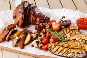Grilled pork knuckle with grilled tomatoes, champignons, vagetable marrow, eggplant, red sweet pepper and baked potatoes and parchment on a wooden cutting board. top view