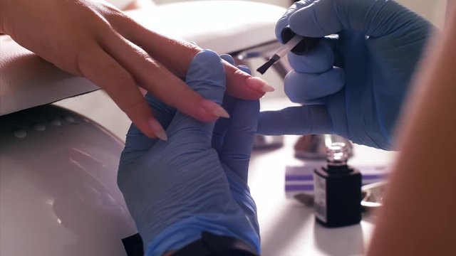 Manicurist master applies a transparent base coat of shellac using brush on client's nail. She is forming client's nail, hands in gloves closeup.
