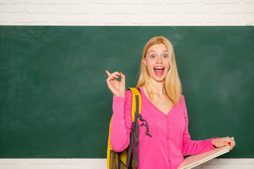 Happy girl holding book and looking at camera while standing in classroom. Student with backpack. Looking for volunteers. High school education. Formal education. Improving myself through education