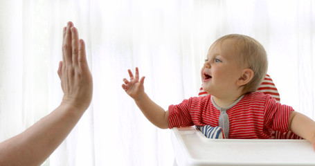 Cute smiling kid sitting on baby chair for feeding and play with faceless parent giving high five