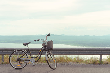 Fototapeta na wymiar Stationary bicycle on cycle path with amazing scenic views of a lake & mountains - Bike with basket parked next to a cliff edge with epic landscape scenery & warm summer filter - image